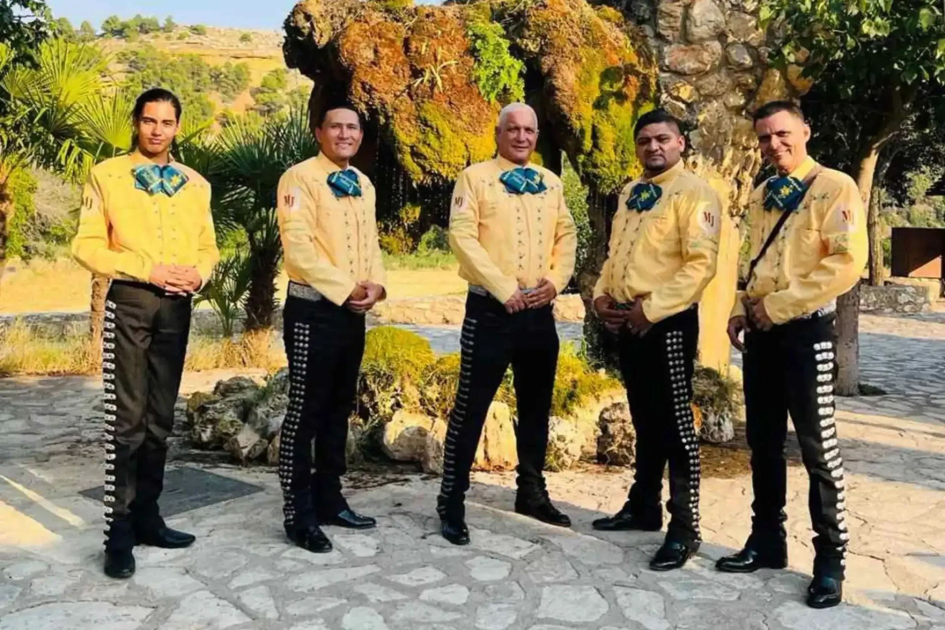 Cinco músicos con chaquetas amarillas y pantalones negros, posando al aire libre.