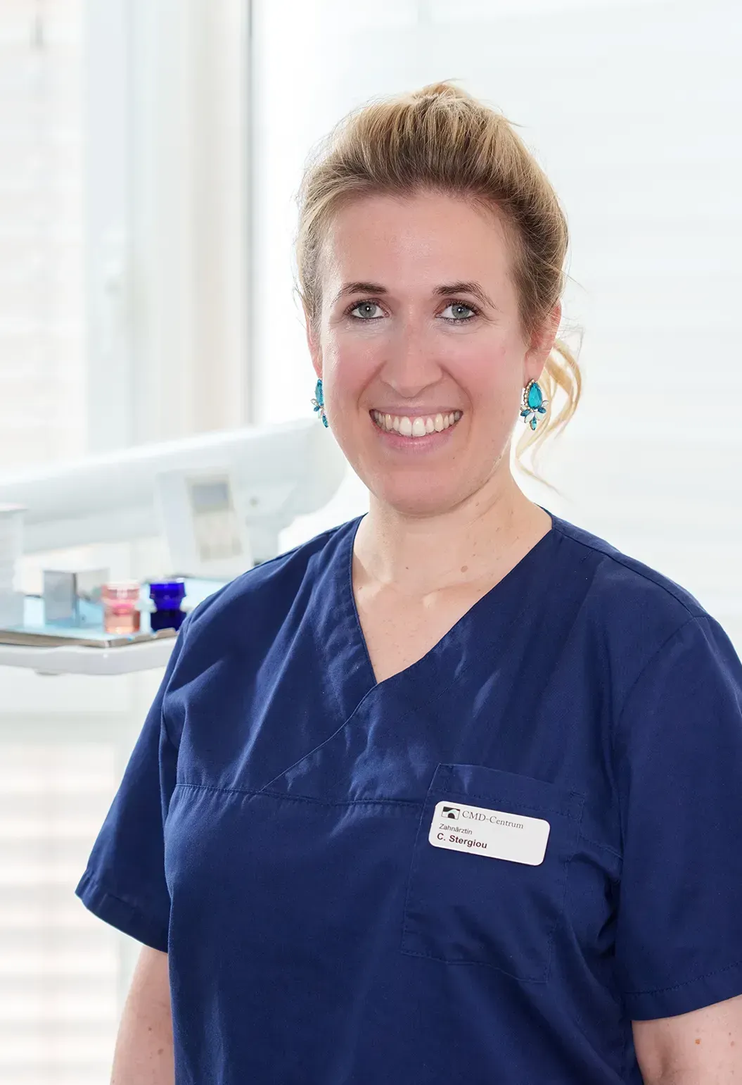 Frau mit heller Haut, grauer OP-Kleidung, lächelt und schaut in die Kamera. Büroumgebung.