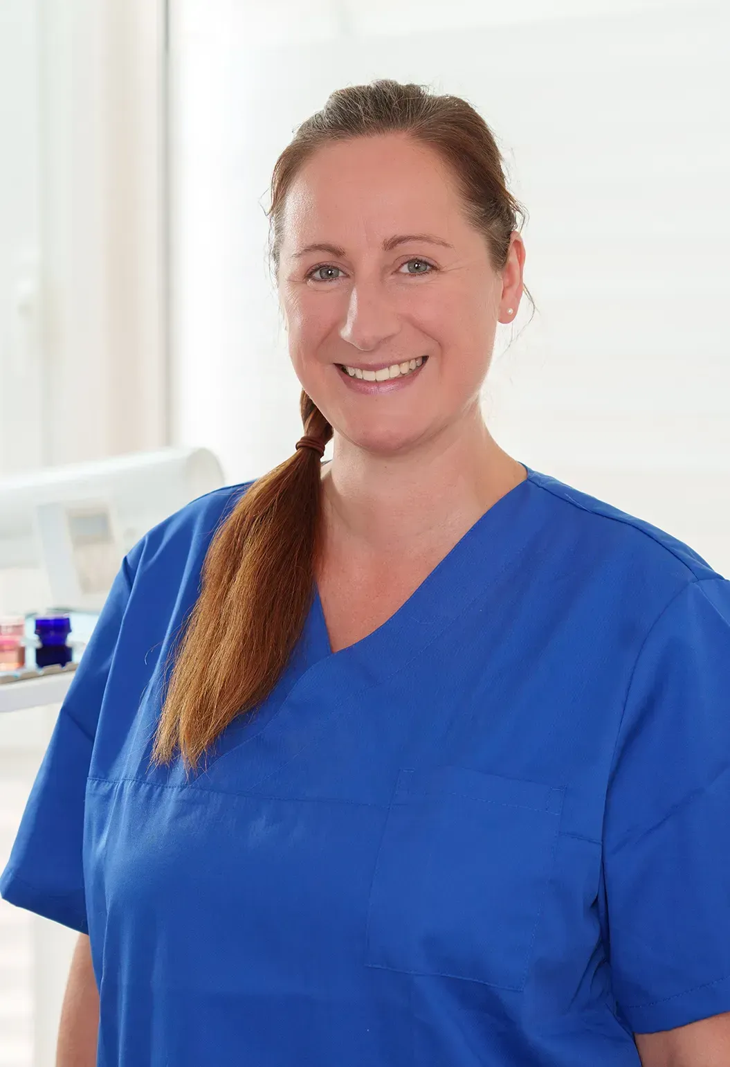 Frau im blauen OP-Kittel lächelt; sanfte Beleuchtung in einem medizinischen Umfeld.