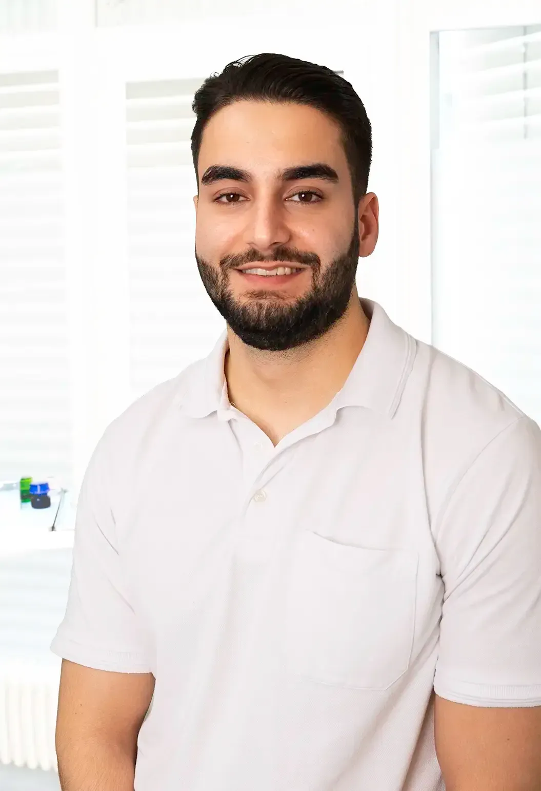 Frau mit heller Haut, grauer OP-Kleidung, lächelt und schaut in die Kamera. Büroumgebung.