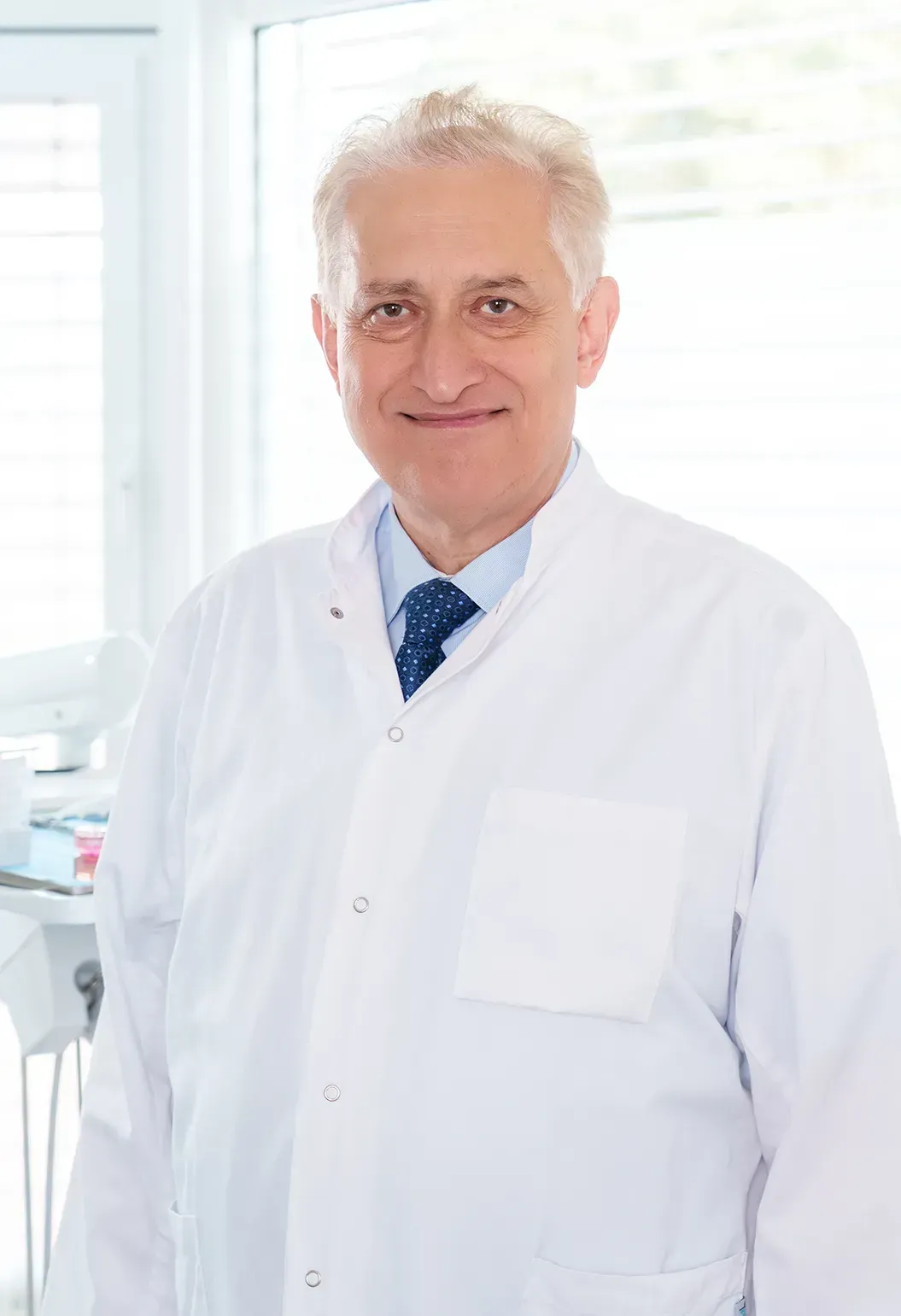 Ein Mann in weißem Mantel und Krawatte steht vor einem Fenster.