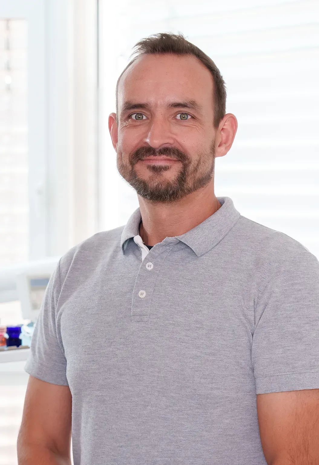 Mann mit kurzen braunen Haaren und Bart, lächelnd, trägt ein graues Poloshirt.
