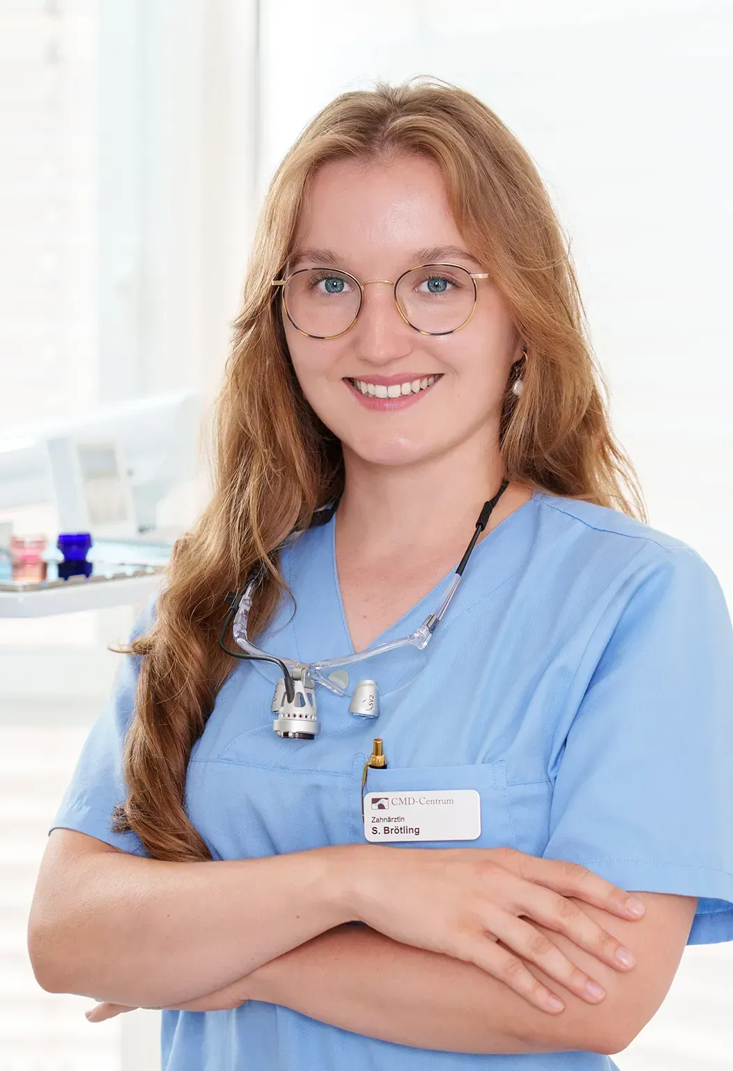Junge Zahnärztin im OP-Kittel, lächelnd mit Brille und Lupenbrille, in einer Praxis.