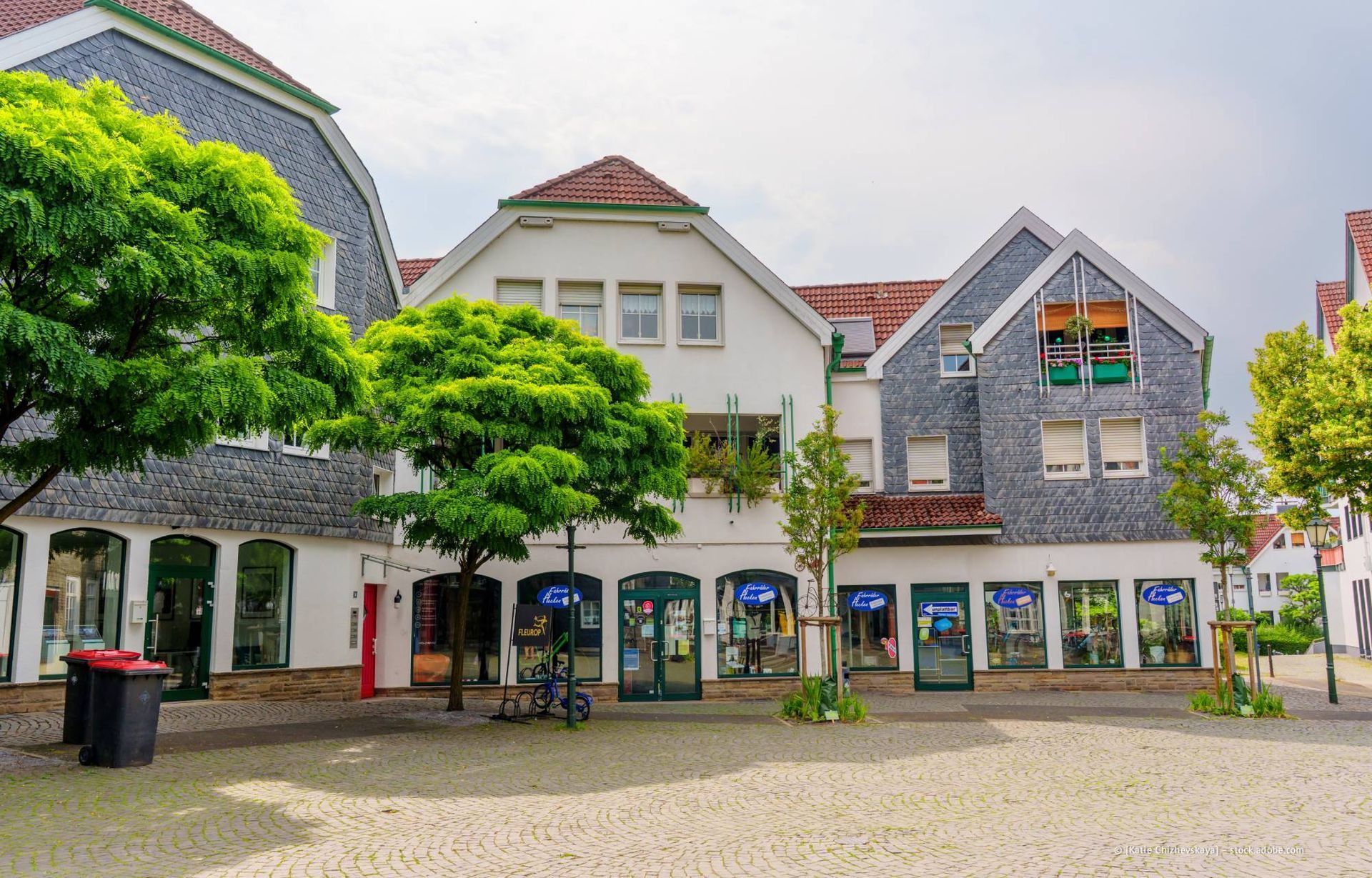 Geschäfte auf einem europäischen Stadtplatz unter Bäumen und bewölktem Himmel.