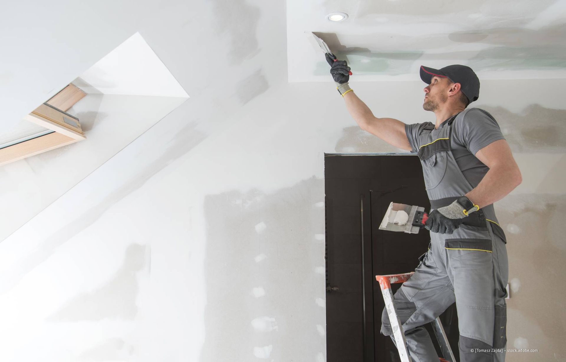 Mann auf Leiter trägt Spachtelmasse an der Decke auf.