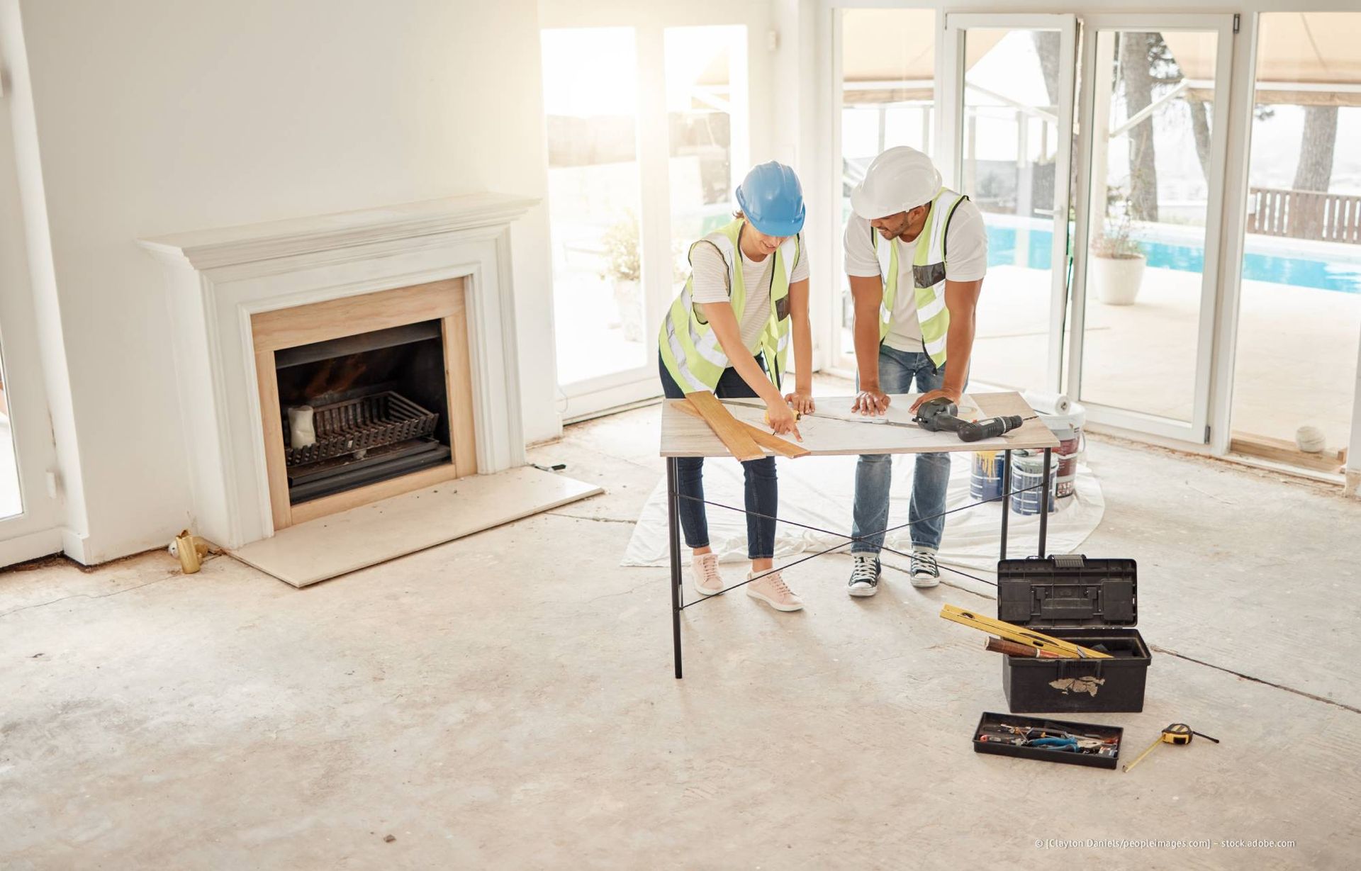 Zwei Bauarbeiter planen ihre Arbeit an einem Arbeitstisch in einem Raum mit Kamin und Blick auf den Pool.