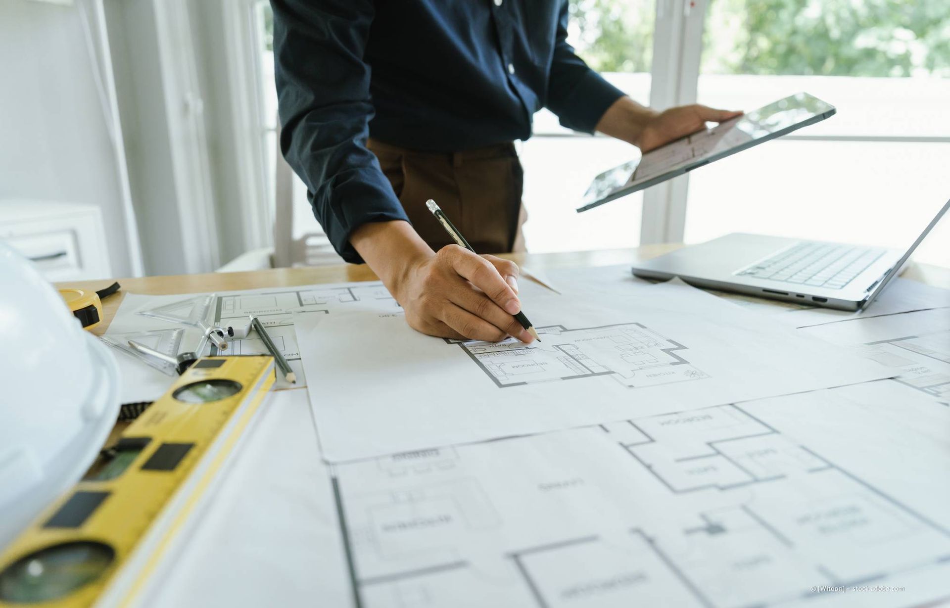 Ein Architekt skizziert in einem Innenraum an einem Schreibtisch mit Laptop, Tablet und Werkzeugen Baupläne.