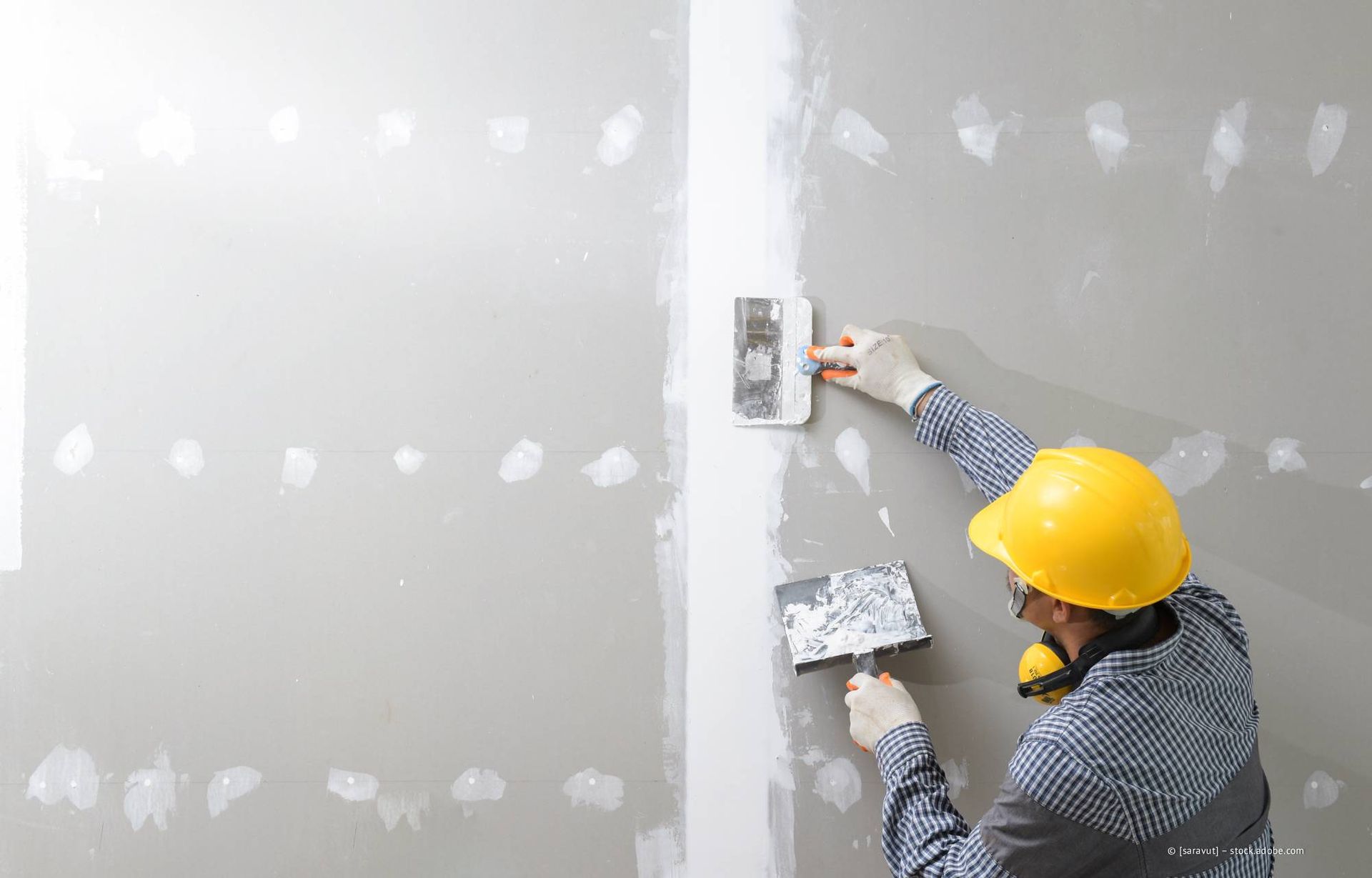 Person mit gelbem Schutzhelm trägt Spachtelmasse auf eine Wand auf.