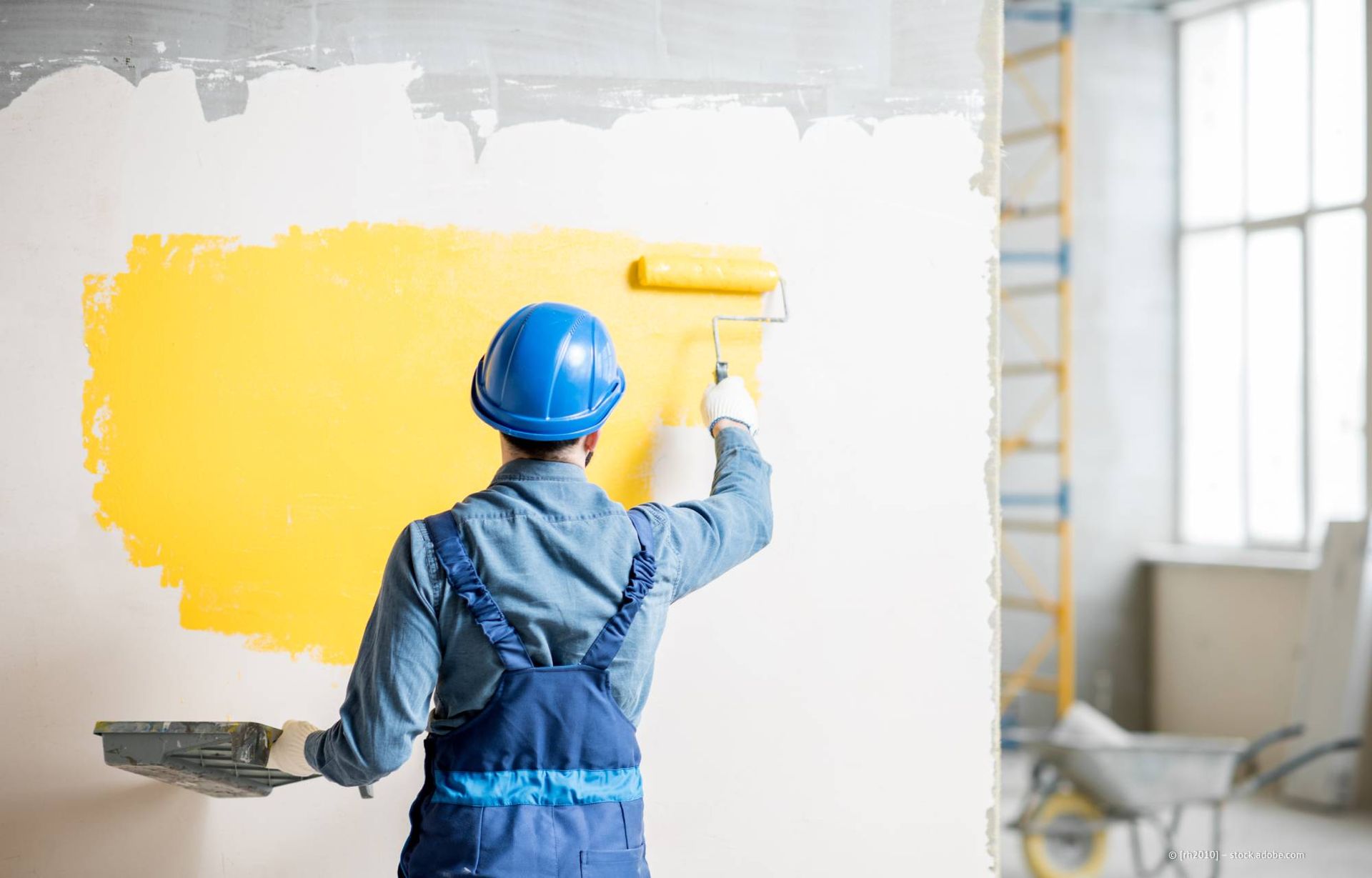 Ein Bauarbeiter in blauem Overall streicht eine Wand mit einer Farbrolle gelb.