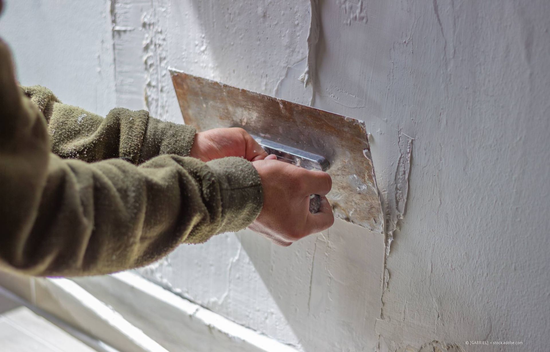 Eine Person trägt mit einer Metallkelle Gips auf eine weiße Wand auf.
