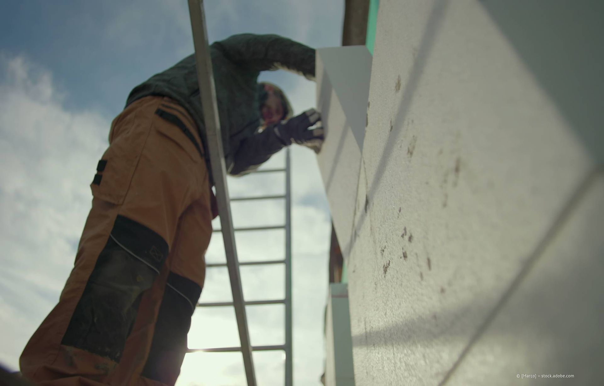 Ein Bauarbeiter auf einer Leiter trägt unter blauem Himmel weiße Farbe auf die Außenfassade eines Gebäudes auf.