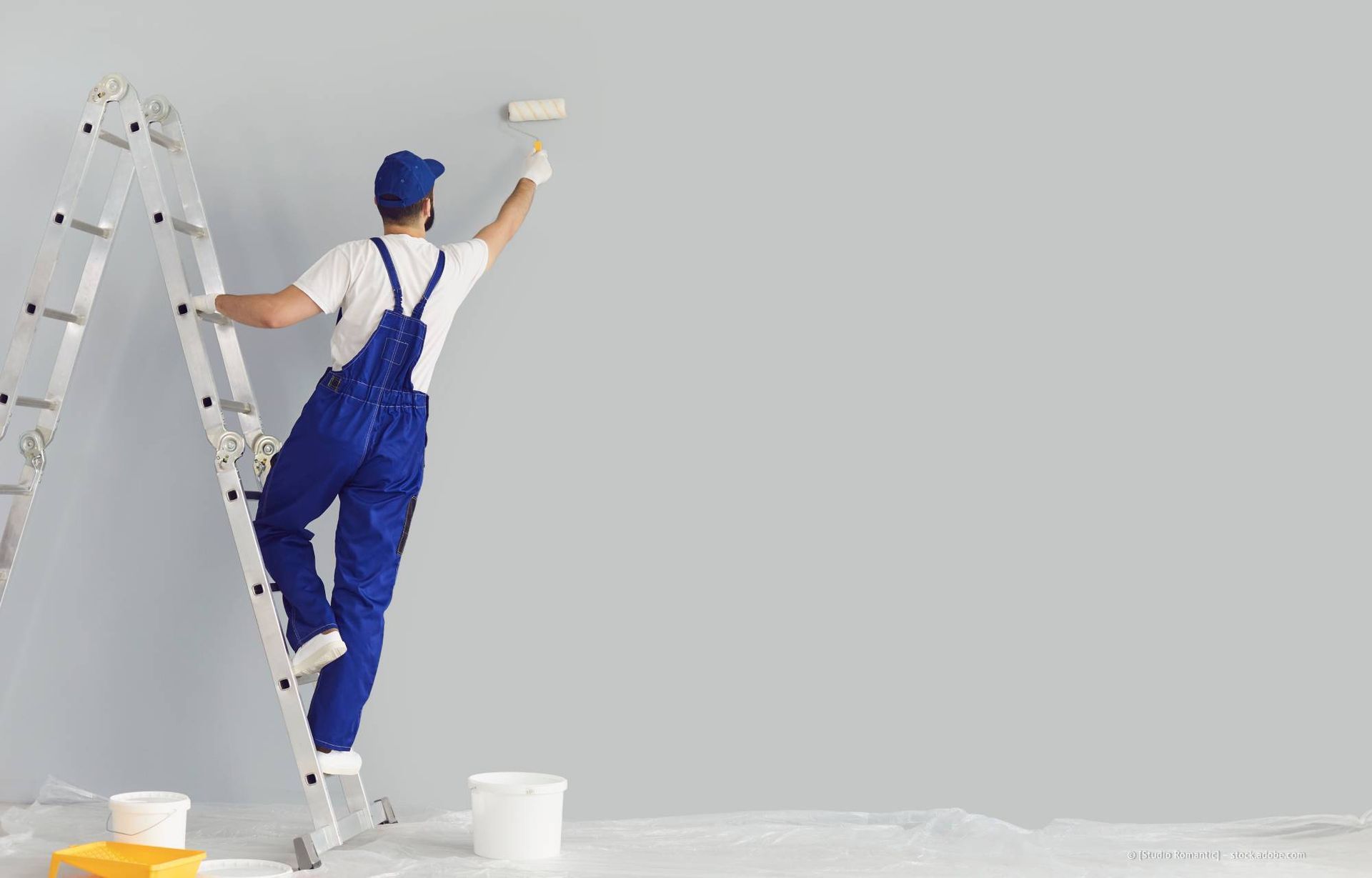 Maler in blauem Overall und mit Kappe auf einer Leiter, der mit einer Farbrolle eine graue Wand streicht.