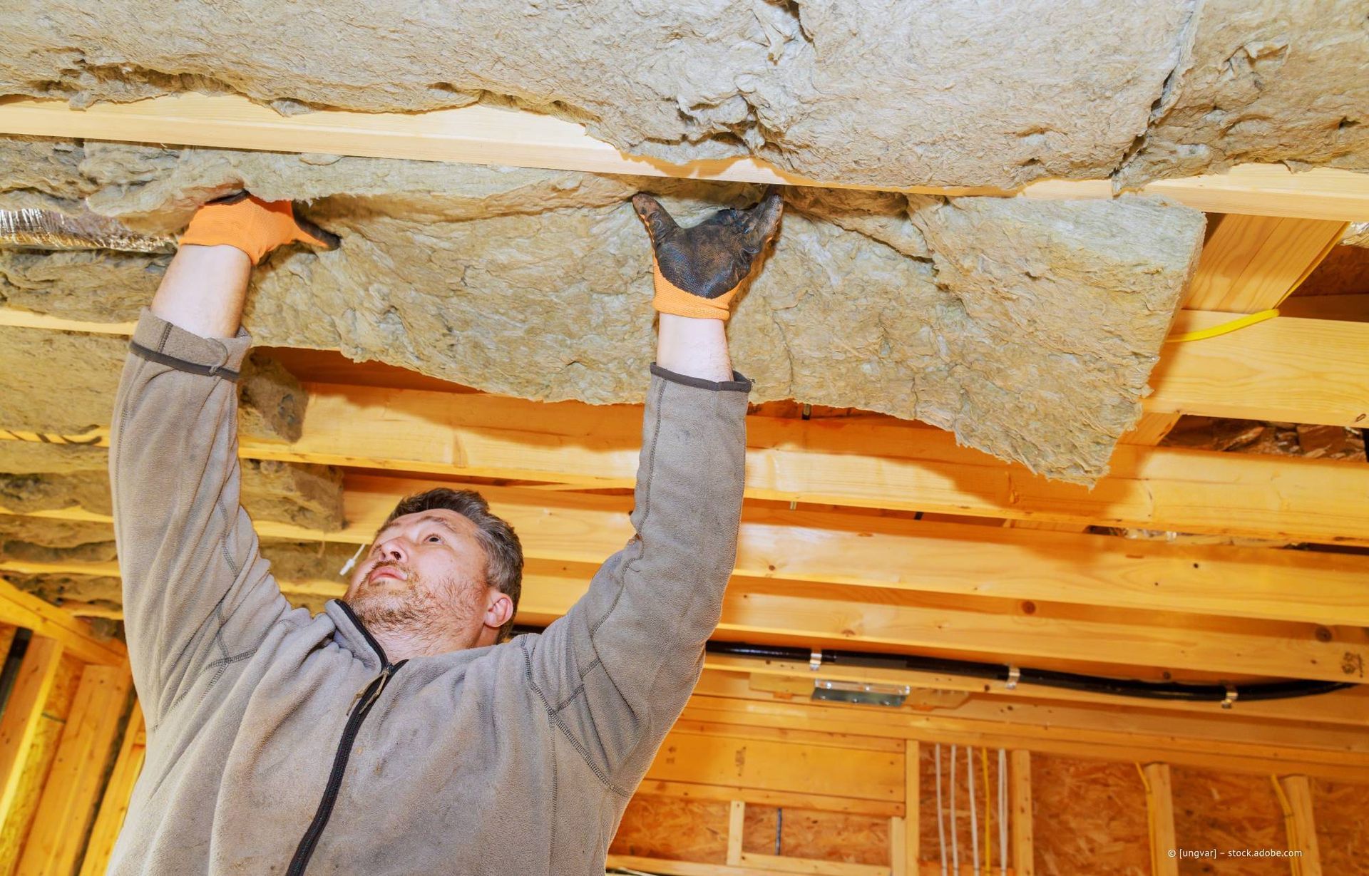 Ein Mann installiert Dämmmaterial in einer Holzrahmendecke. Er trägt Handschuhe und einen grauen Pullover.