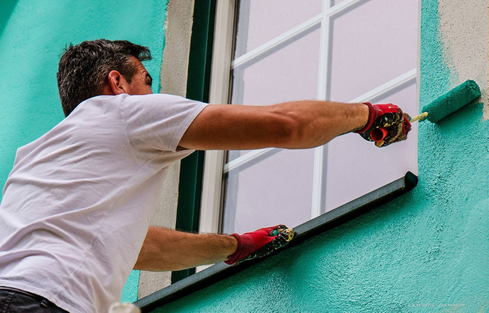 Eine Person streicht die Außenwand um ein Fenster herum mit einer grünen Farbrolle.