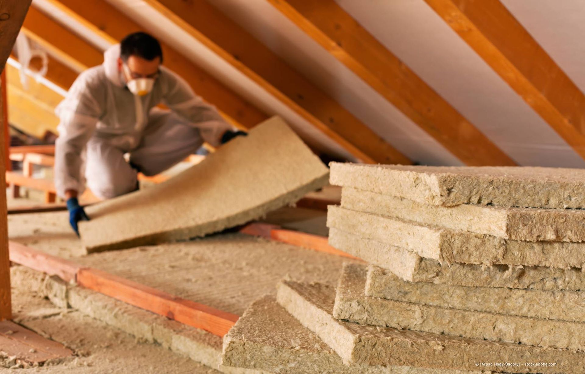 Ein Mann in Schutzkleidung installiert Dämmmaterial auf einem Dachboden und hält dabei eine Dämmplatte neben gestapelten Platten.