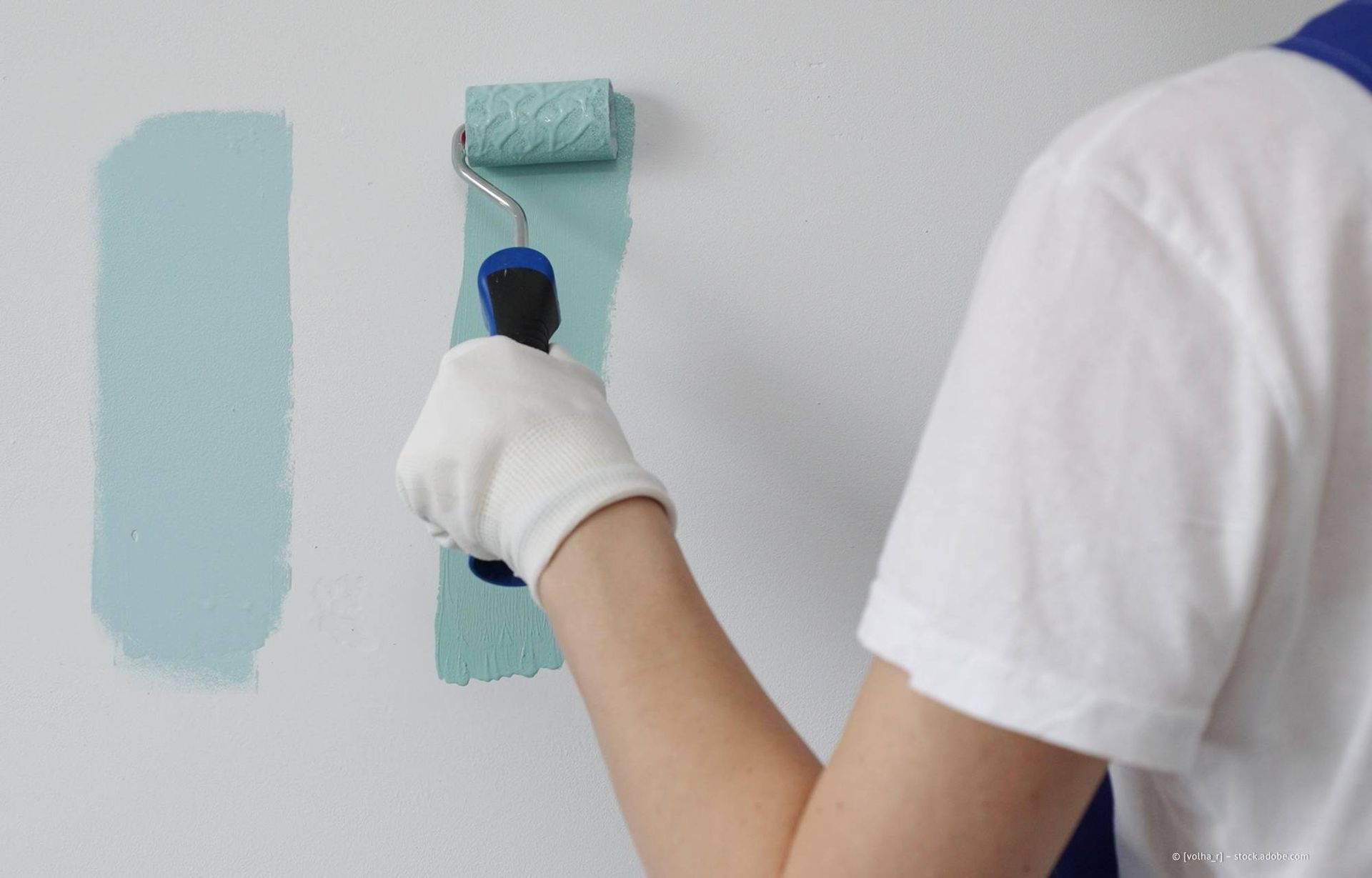 Eine Person in weißem Hemd und Handschuhen malt mit einer Farbrolle einen hellblauen Farbfleck auf eine weiße Wand.