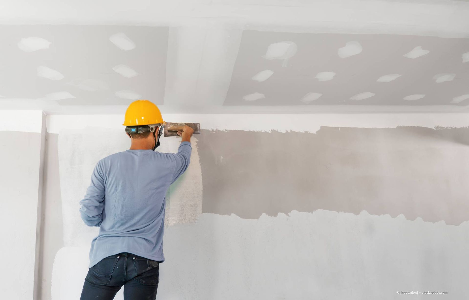 Ein Bauarbeiter mit Schutzhelm glättet Gipskartonplatten an einer Wand.