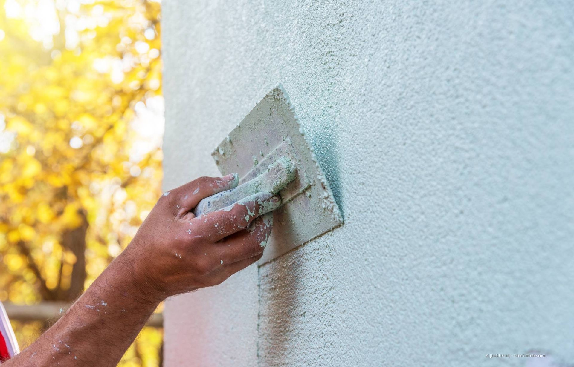 Eine Person trägt mit einer Kelle Stuck auf eine Außenwand auf; sonniger Tag, Laub unscharf.