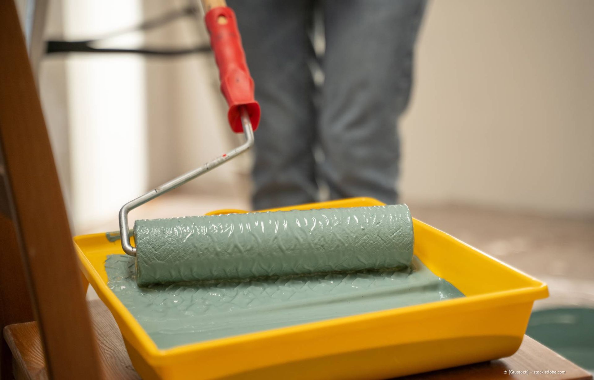 Eine Person streicht eine Wand mit einer Farbrolle in einer Wanne mit hellgrüner Farbe.