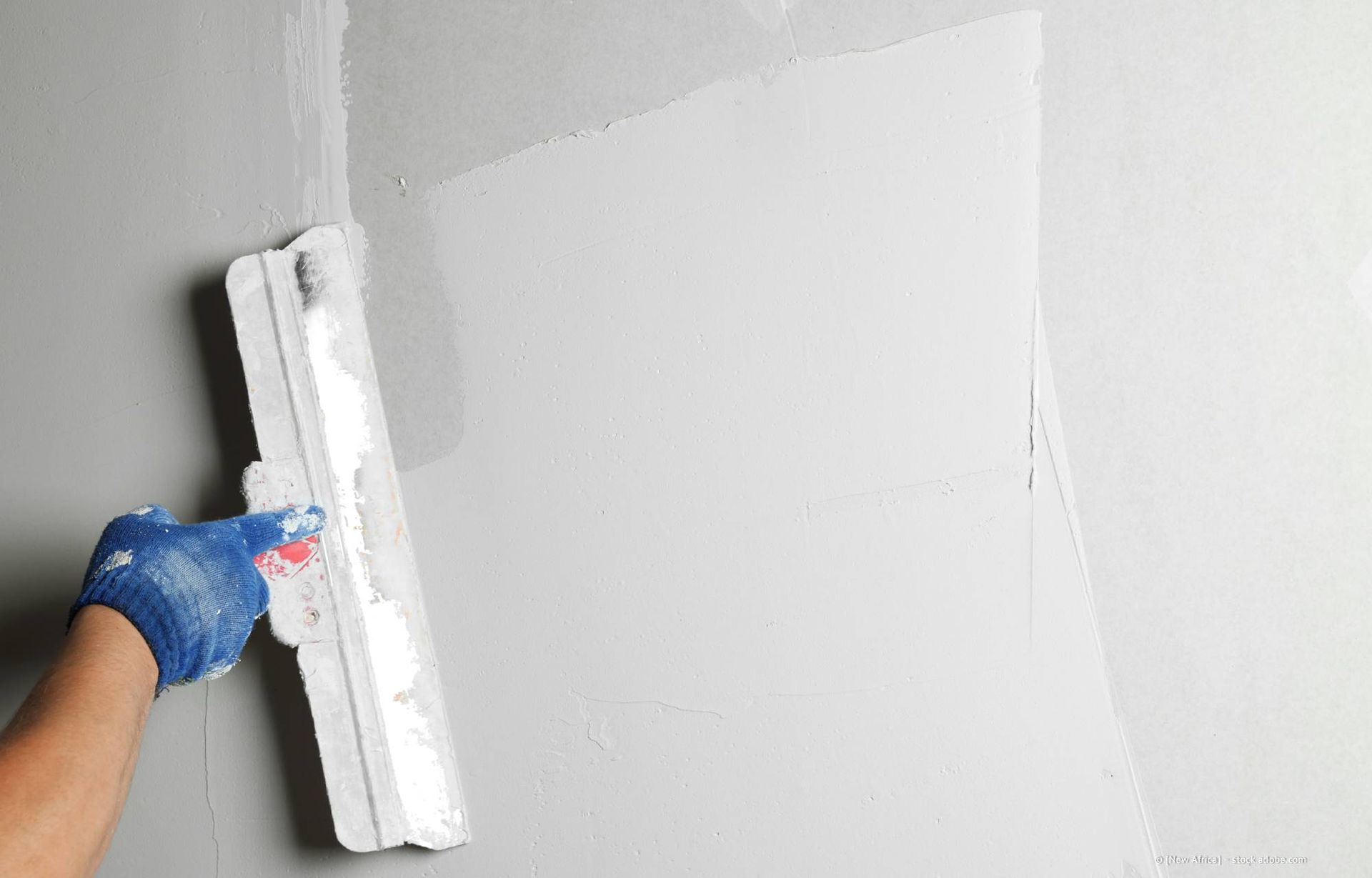 Eine Person, die mit einer Kelle weiße Gipsspachtelmasse auf eine Wand aufträgt, trägt dabei einen blauen Handschuh.