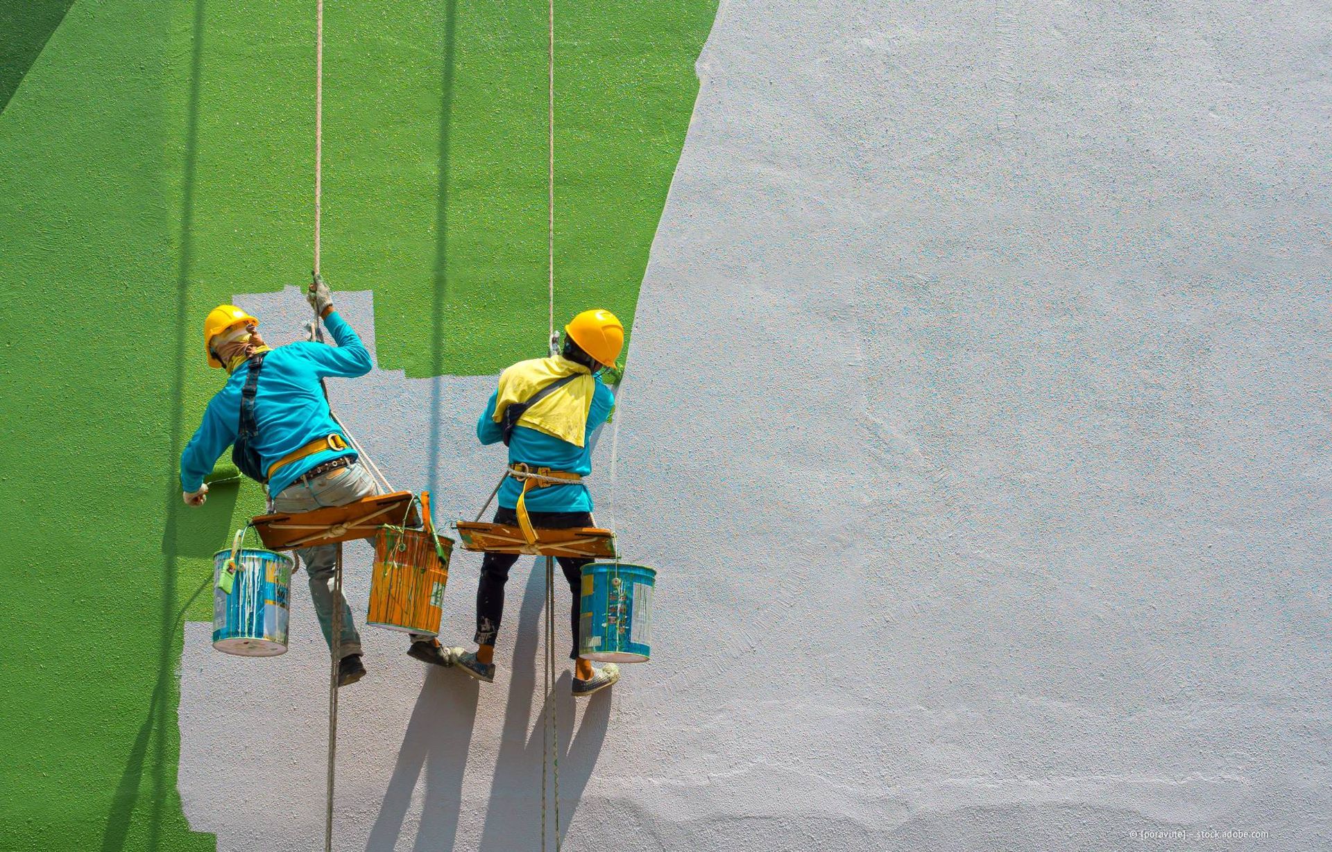 Zwei in Gurten gesicherte Personen streichen, schwebend, eine grüne Wand weiß.