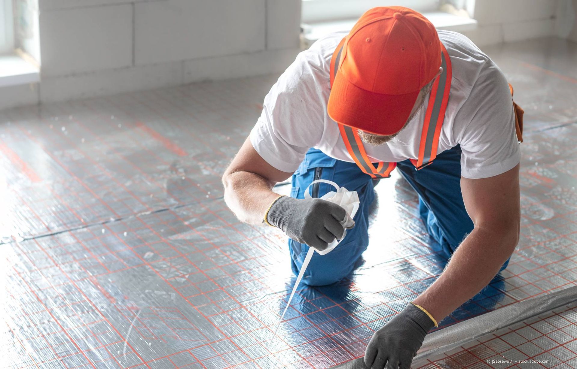 Eine Person, die Bodenbeläge verlegt, trägt eine orangefarbene Kappe und eine Sicherheitsweste und trägt Dichtmittel auf.