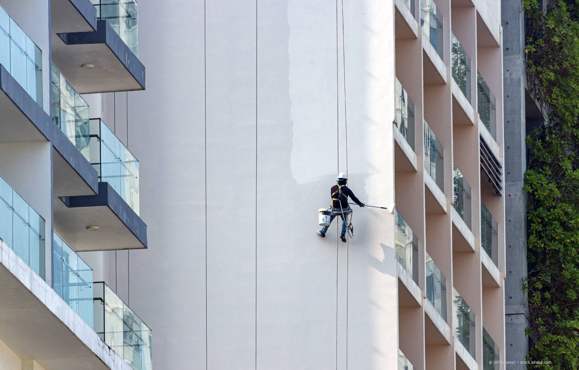 Ein Maler an Seilen trägt weiße Farbe auf die Wand eines Hochhauses auf.