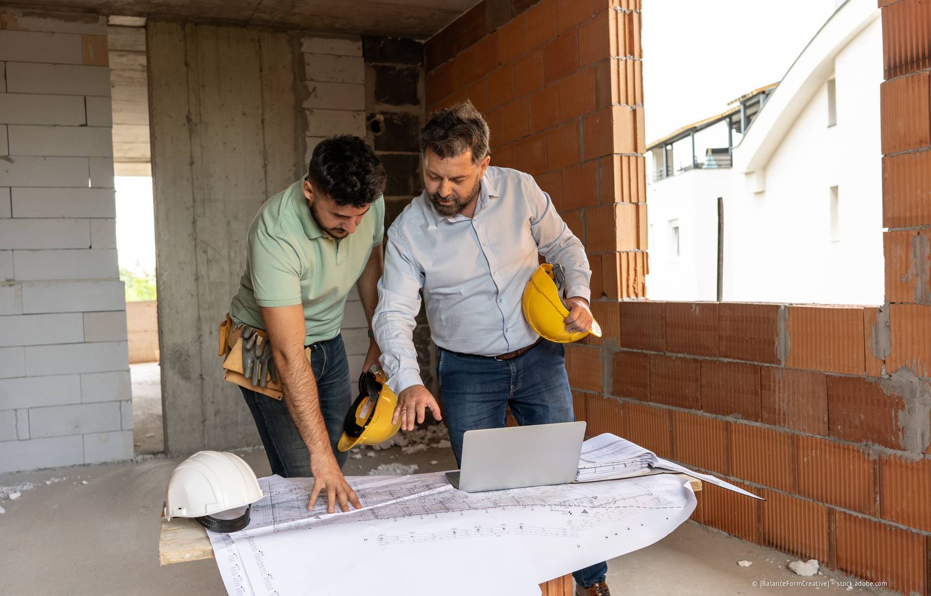 Zwei Männer studieren Baupläne auf einer Baustelle, einer mit einem Laptop, der andere mit einem Schutzhelm.
