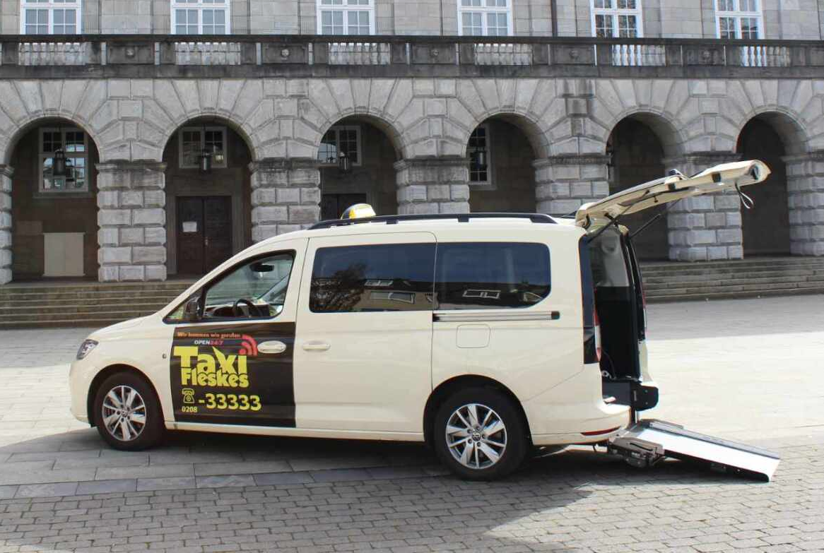 Barrierefreies Taxi mit ausgefahrener Rampe, geparkt vor einem Gebäude mit gewölbten Eingängen.