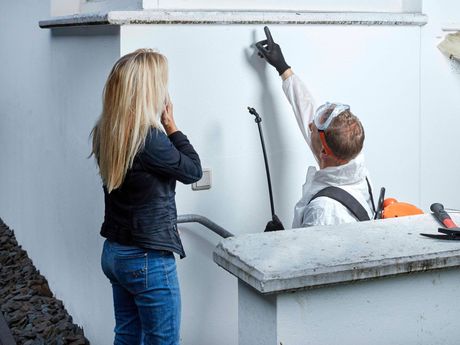 Frau beobachtet einen Mann in Schutzkleidung, der auf eine weiße Wand zeigt, Sprühgerät in der Nähe. Im Freien.