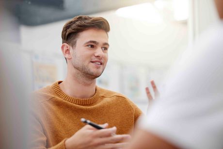 Mann im orangefarbenen Pullover gestikuliert beim Sprechen, hält Stift, drinnen.