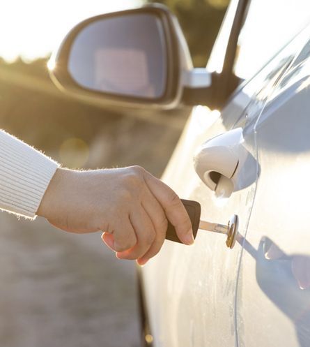 Una mano gira la llave de la puerta del coche en la cerradura, con la luz del sol reflejándose en el vehículo blanco.