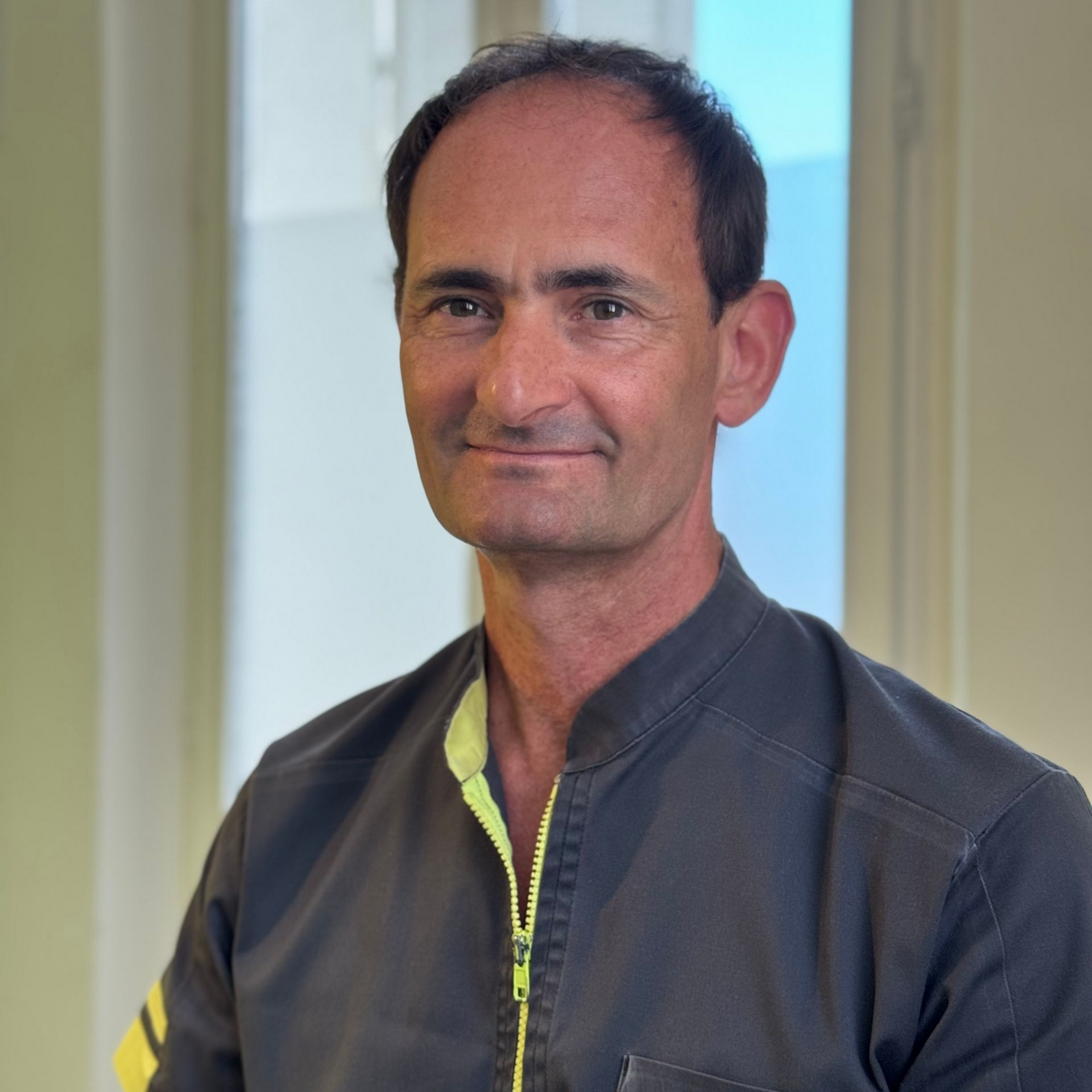 Homme en uniforme gris à fermeture éclair jaune, souriant, à l'intérieur.