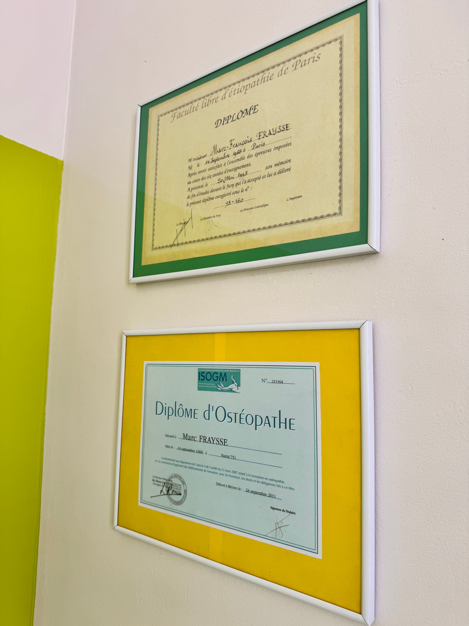 Deux certificats encadrés sont accrochés à un mur blanc ; l'un avec un cadre vert, l'autre avec un cadre jaune.