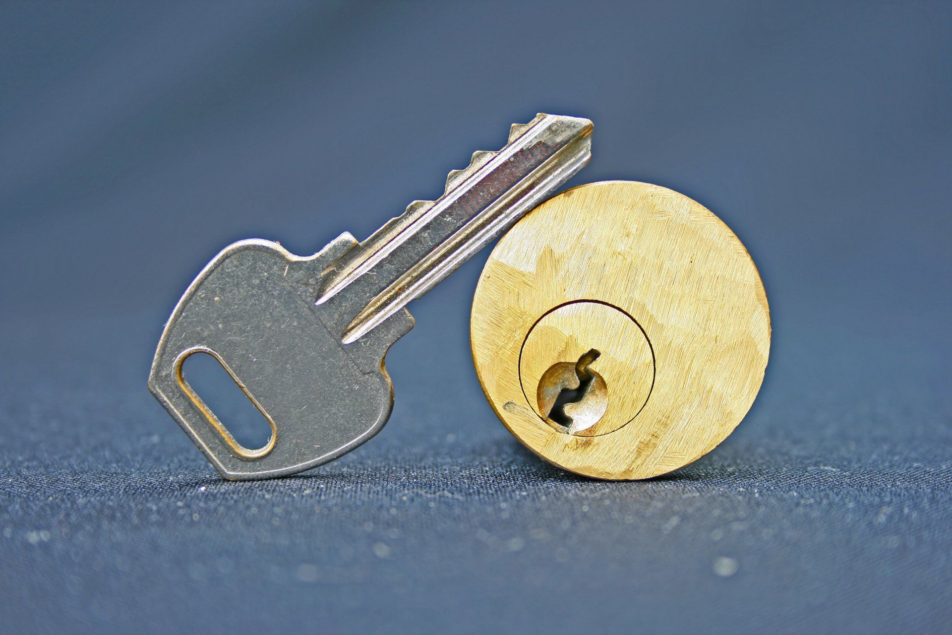 Un cylindre de serrure circulaire couleur laiton repose à côté d'une clé en métal argenté sur un fond bleu foncé.