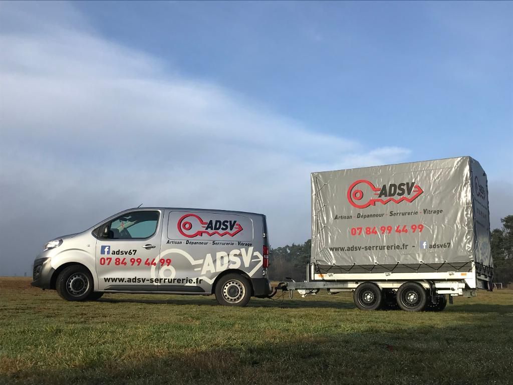 Une camionnette argentée et une remorque assortie, portant le logo et les coordonnées d'ADSV, sont garées sur un terrain herbeux.