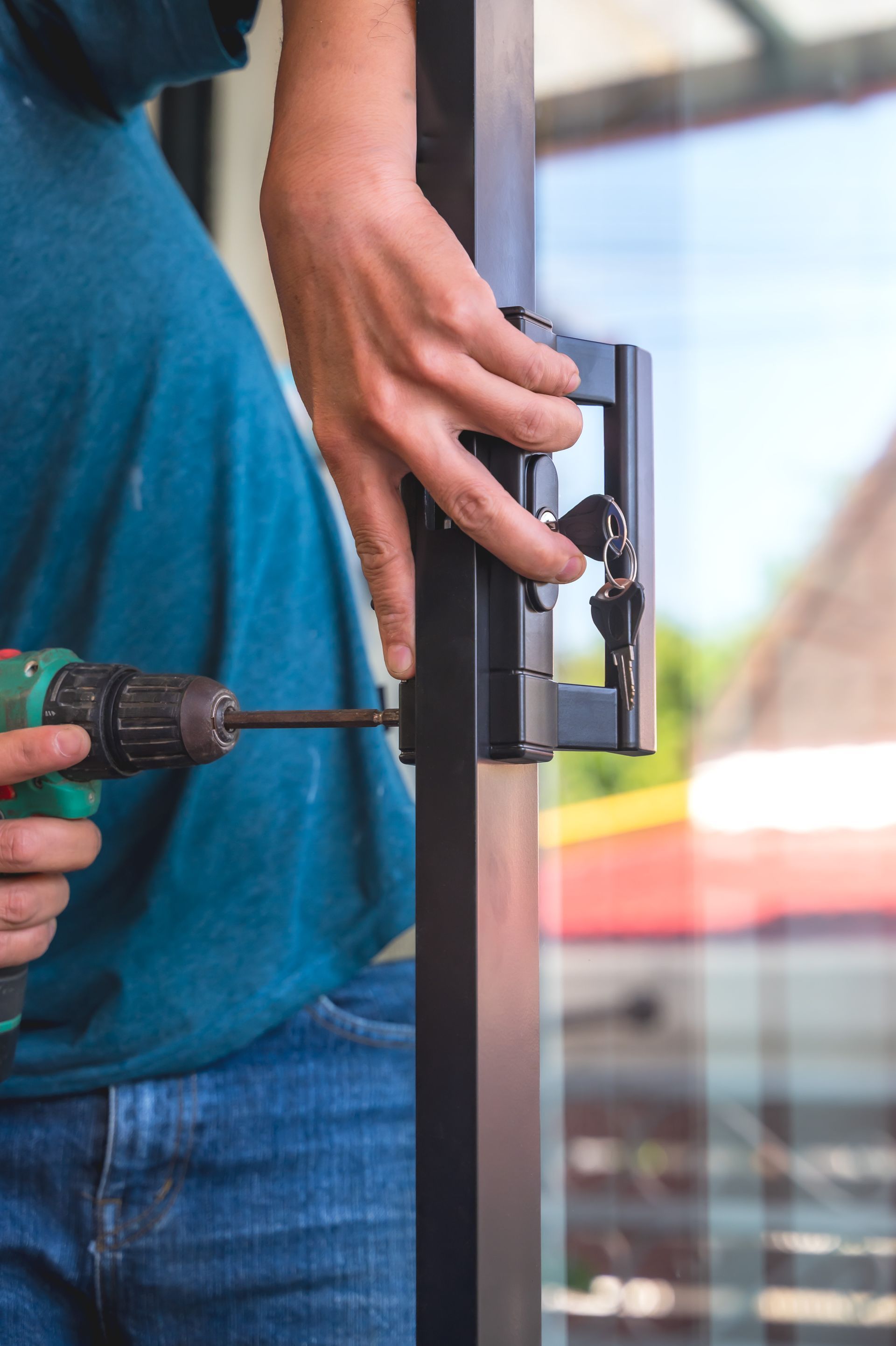 Une personne vêtue d'une chemise bleue utilise une perceuse sans fil pour installer une poignée de porte noire sur un cadre de porte vitrée.