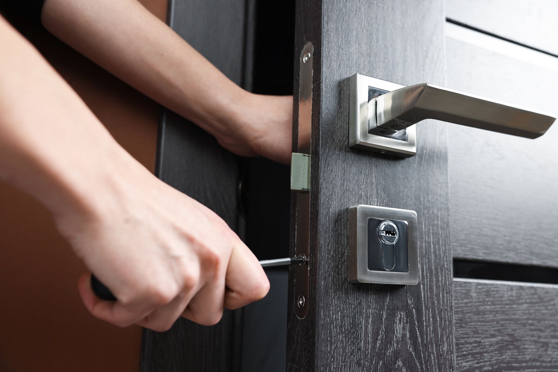 Une main utilise un tournevis pour réparer le mécanisme de verrouillage d'une porte gris foncé.