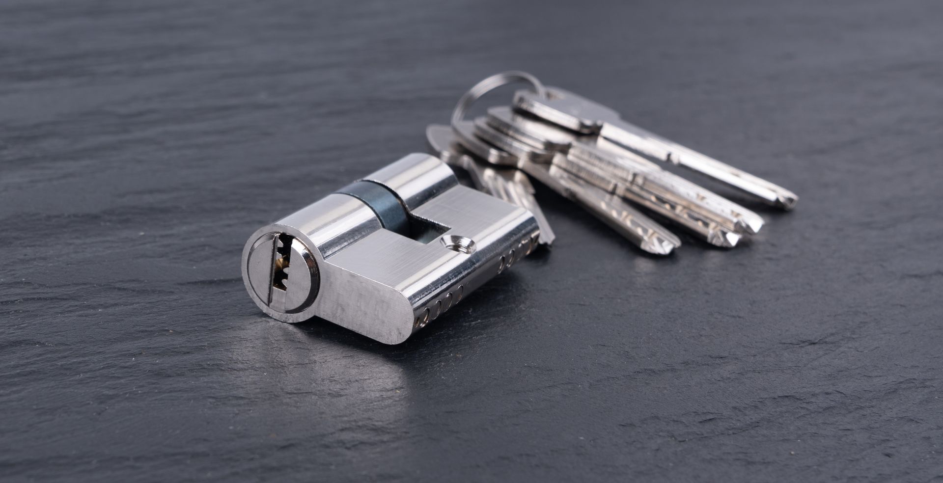 Un cylindre de serrure de porte argenté et un trousseau de clés posés sur une surface sombre et texturée.