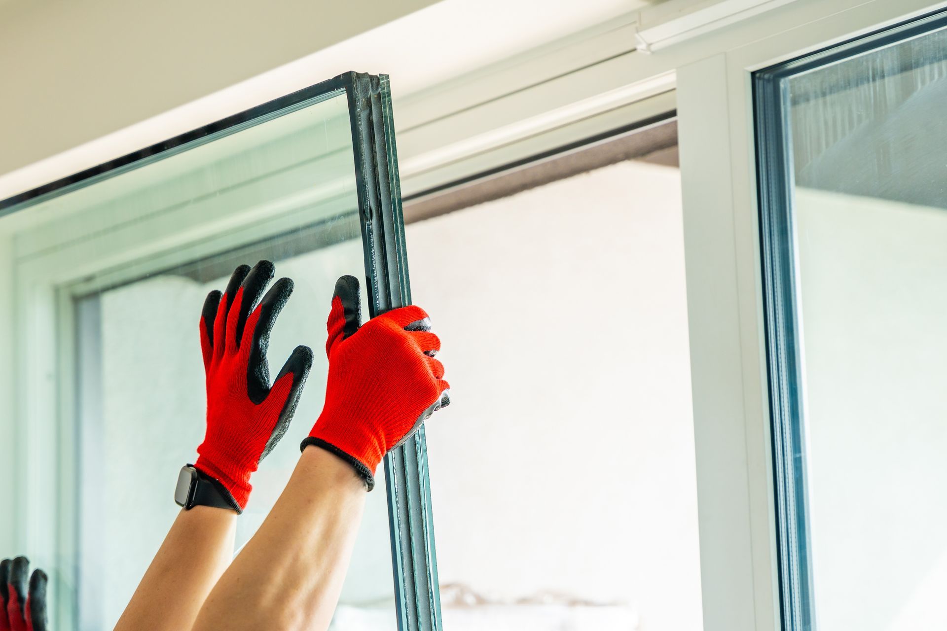 Deux mains gantées de gants de travail rouges et noirs installent soigneusement une vitre dans un cadre de fenêtre.