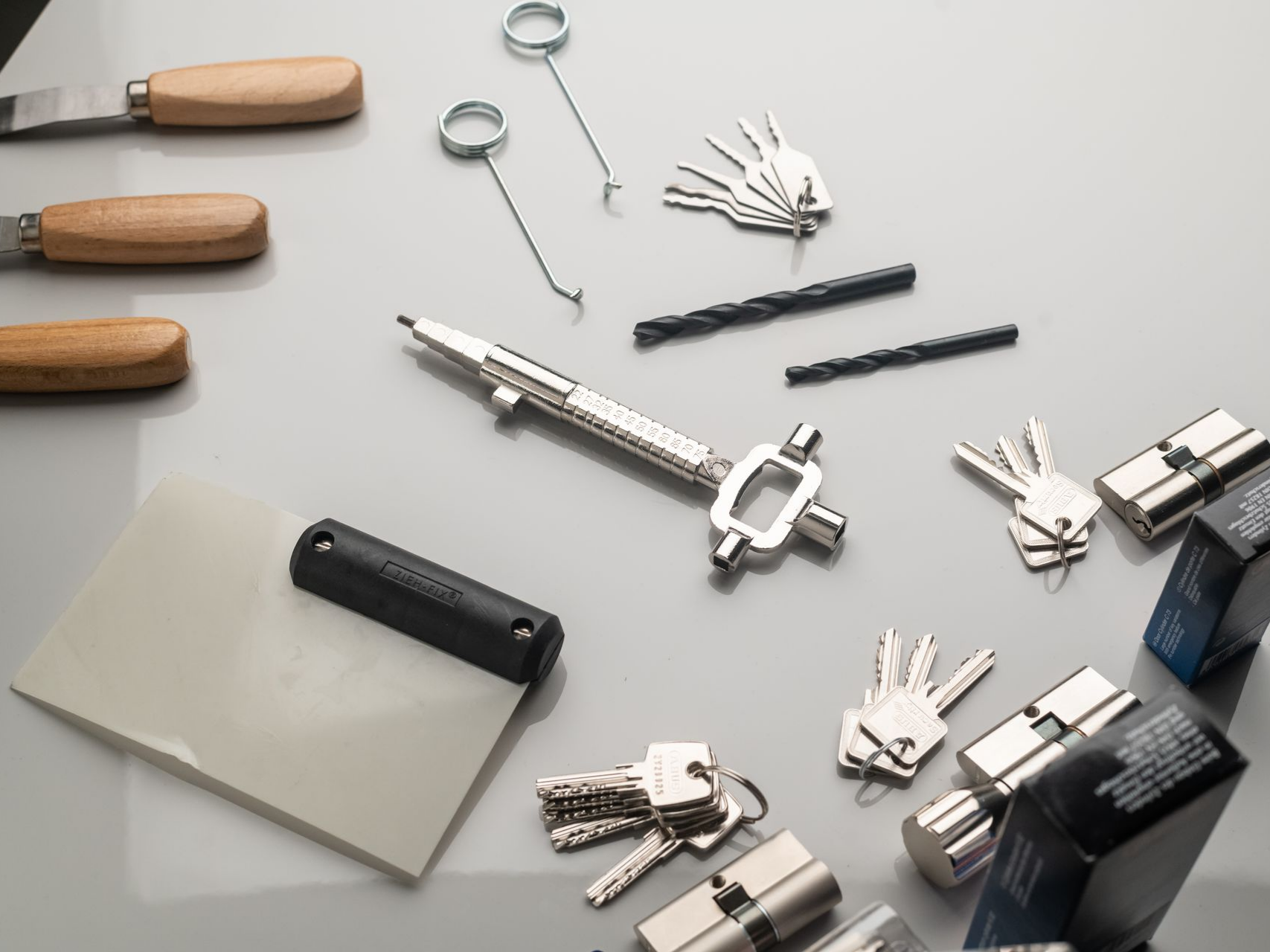 Outils de serrurier, notamment des serrures à cylindre, des clés, des clés de tension, un gabarit de clé, des perceuses et des couteaux à mastic, sur une surface blanche.
