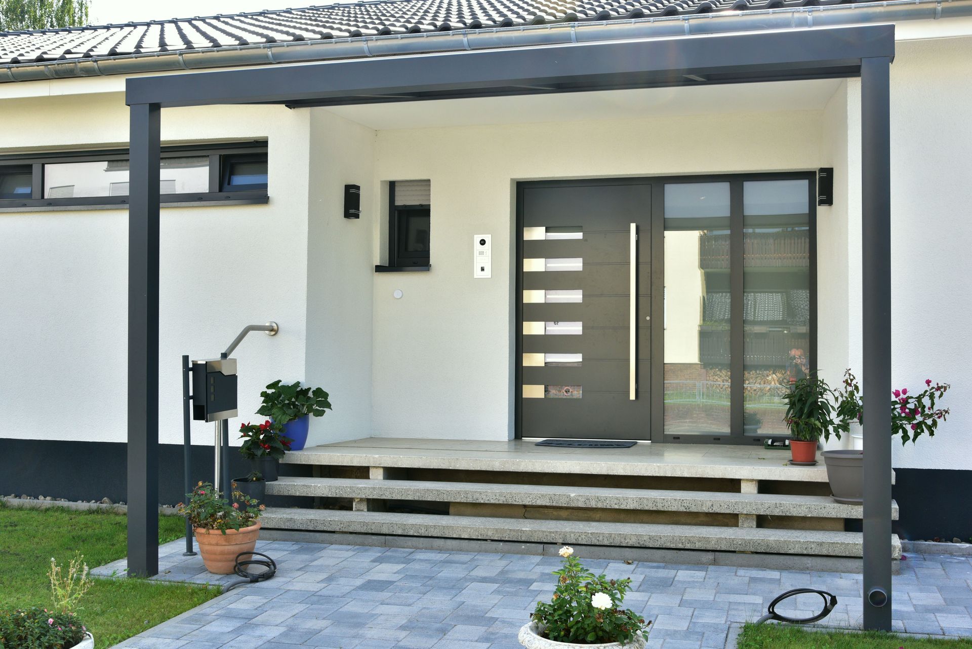 Entrée moderne avec une porte grise, trois marches en pierre, une structure en métal foncé et plusieurs plantes en pot.