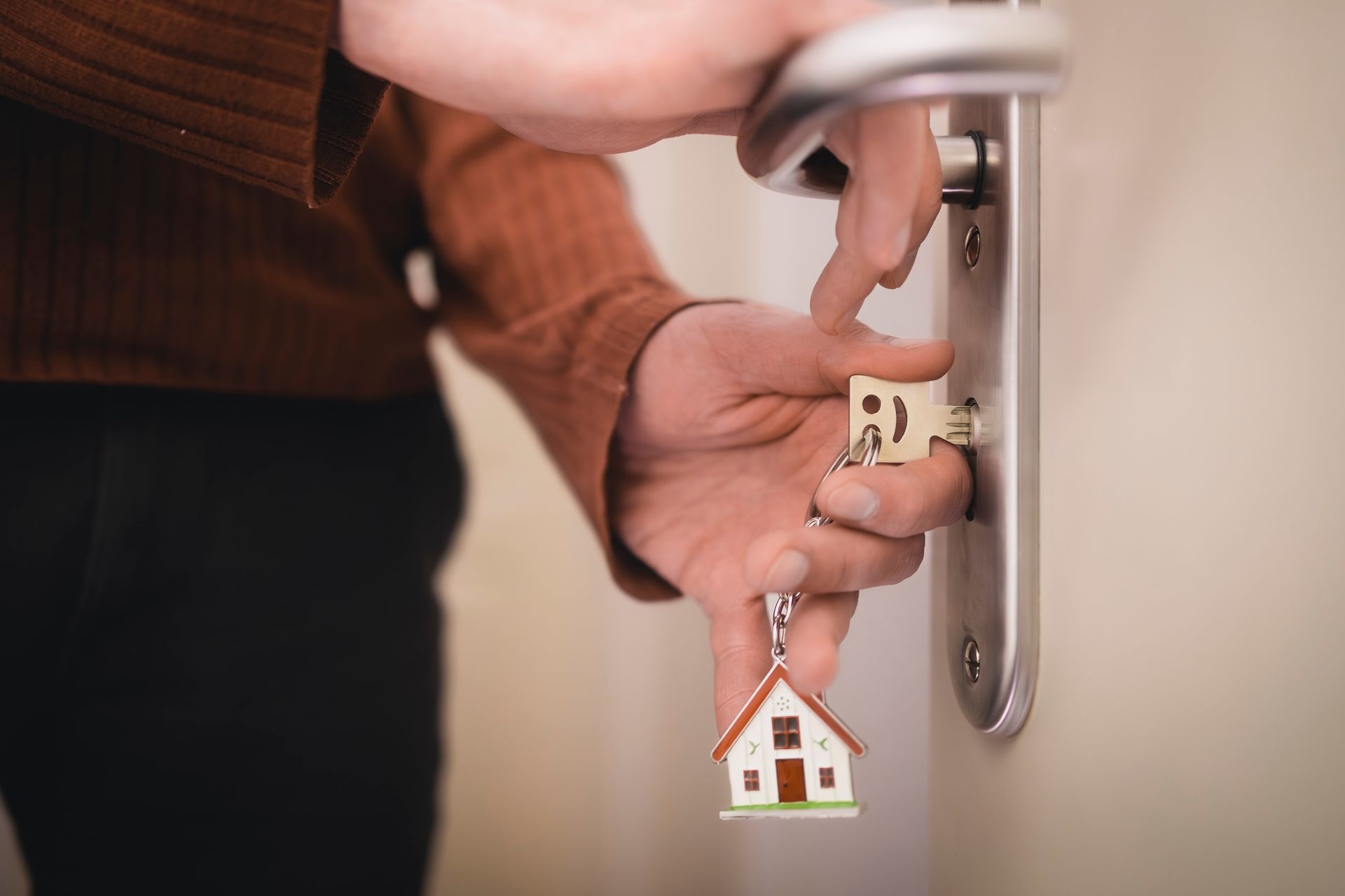 Une personne portant un pull marron insère une clé munie d'un petit porte-clés en forme de maison dans une serrure de porte argentée.