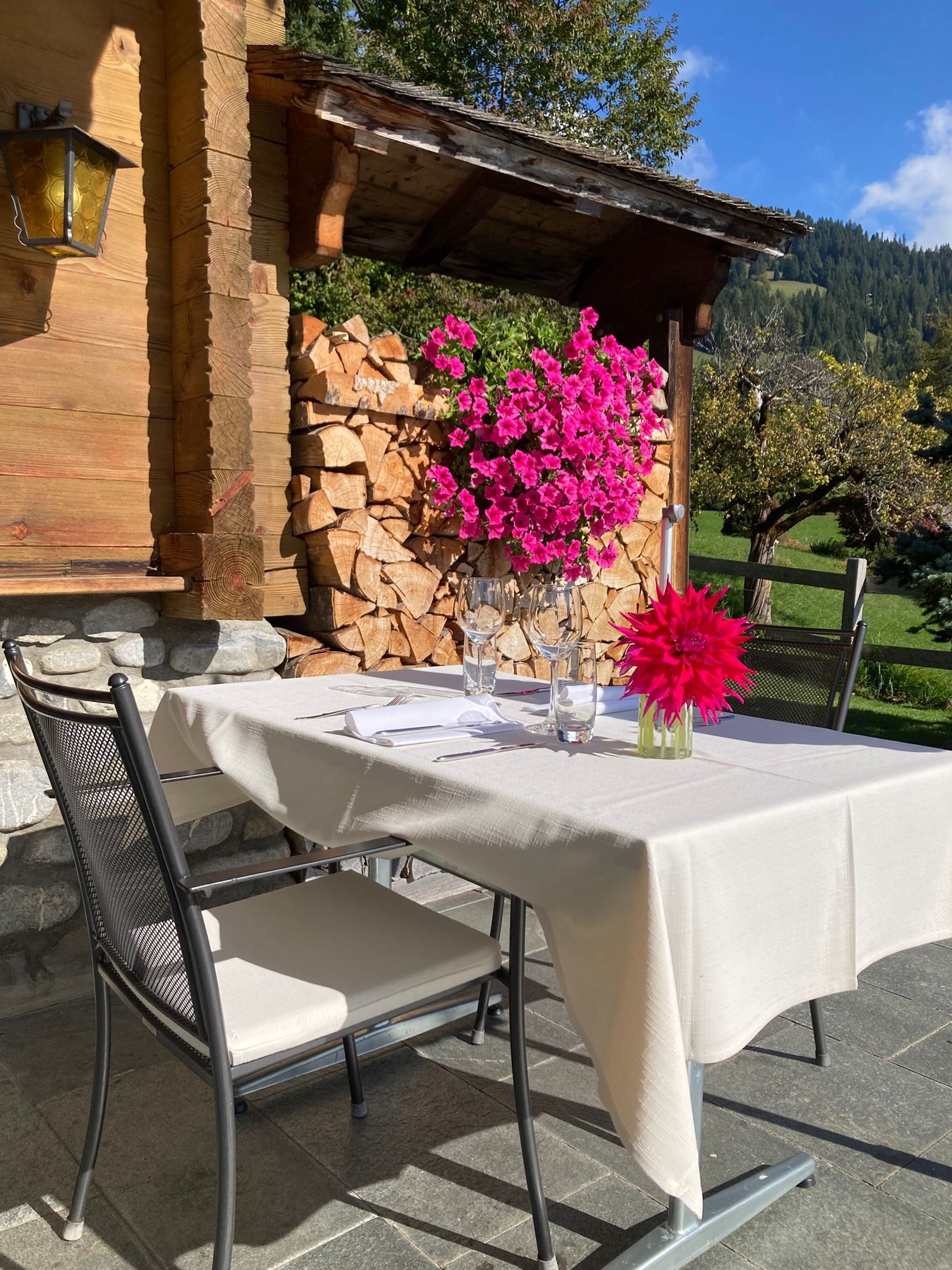 Ein Tisch mit leuchtend pinkfarbenen Blumen auf einer Terrasse neben einer rustikalen, mit Baumstämmen gefüllten Mauer.