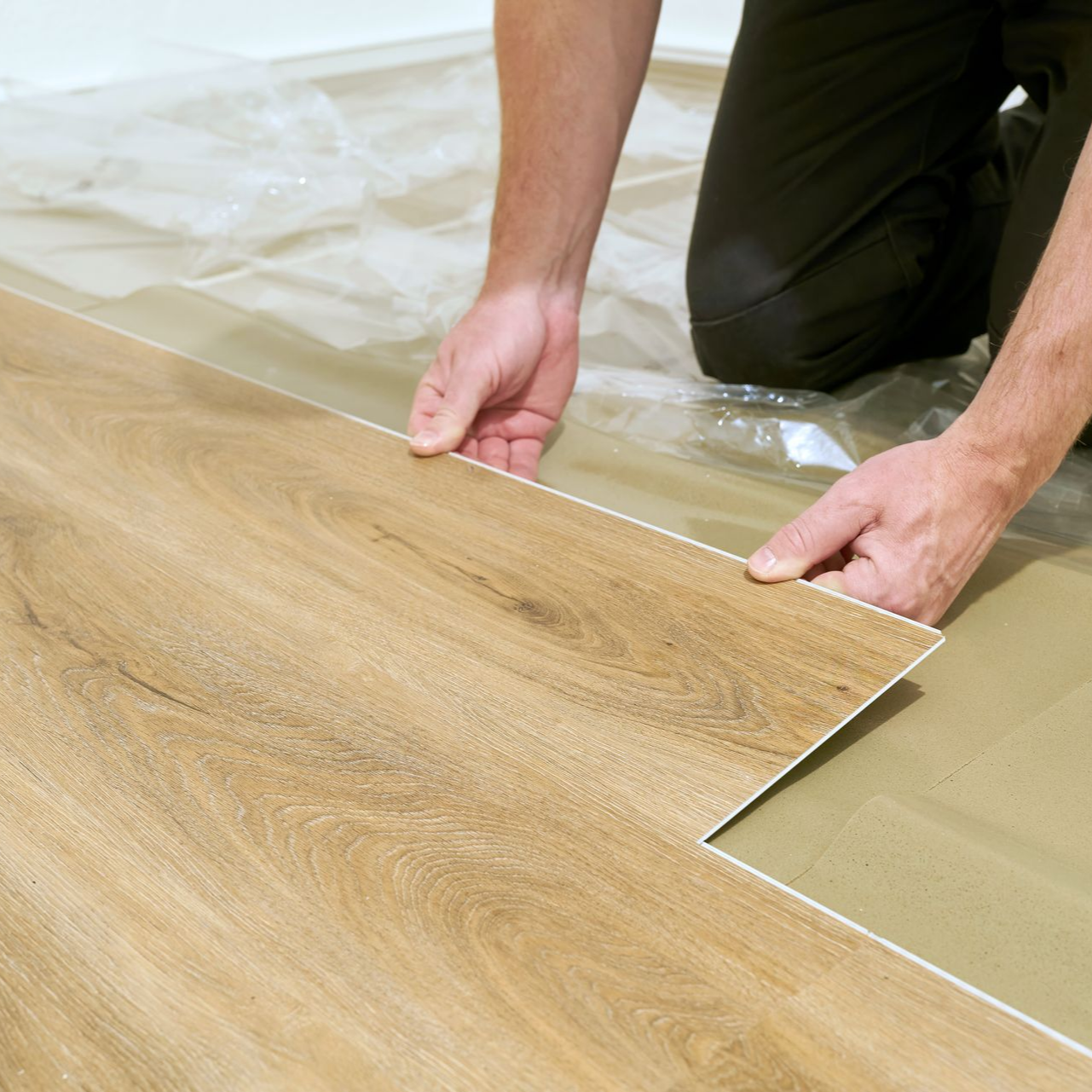 Personne posant un revêtement de sol imitation bois, agenouillée et tenant une lame près de lames déjà posées.