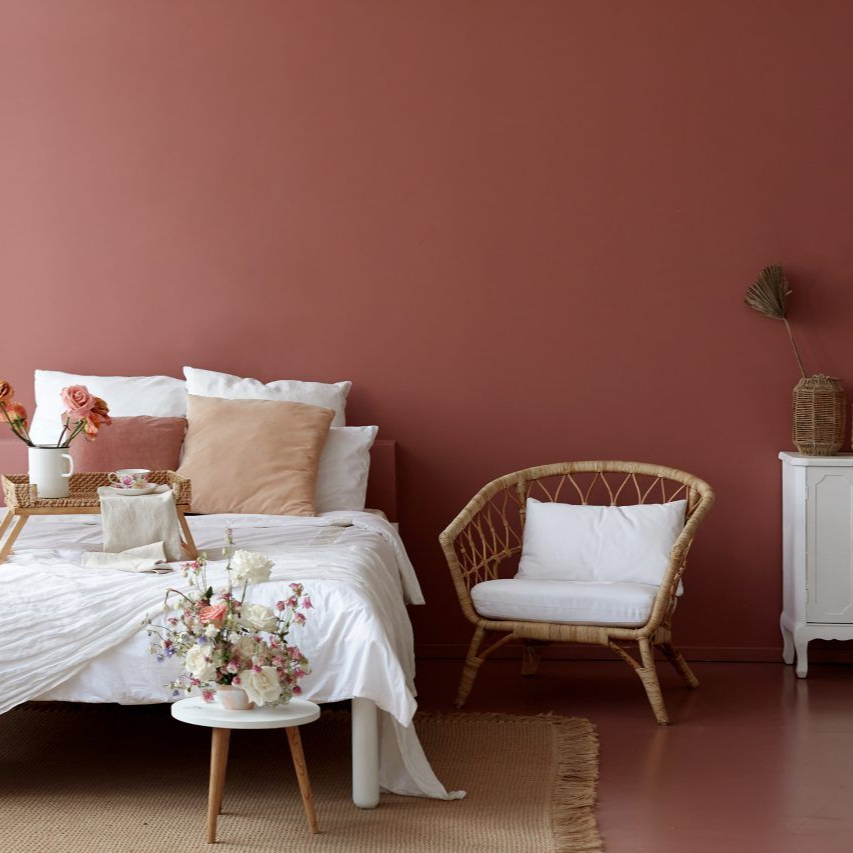 Chambre avec lit, fauteuil en rotin, table de chevet blanche et murs brun rougeâtre.