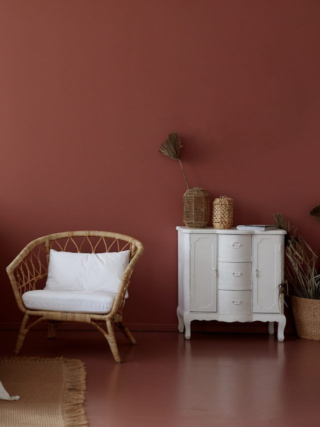 Fauteuil en rotin avec coussin blanc, armoire blanche, objets décoratifs sur un mur et un sol rouge foncé.