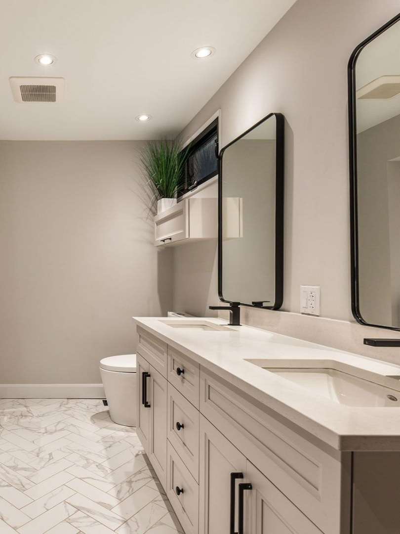 Salle de bains moderne avec meubles blancs, sol en marbre, miroirs à cadre noir et une petite fenêtre.