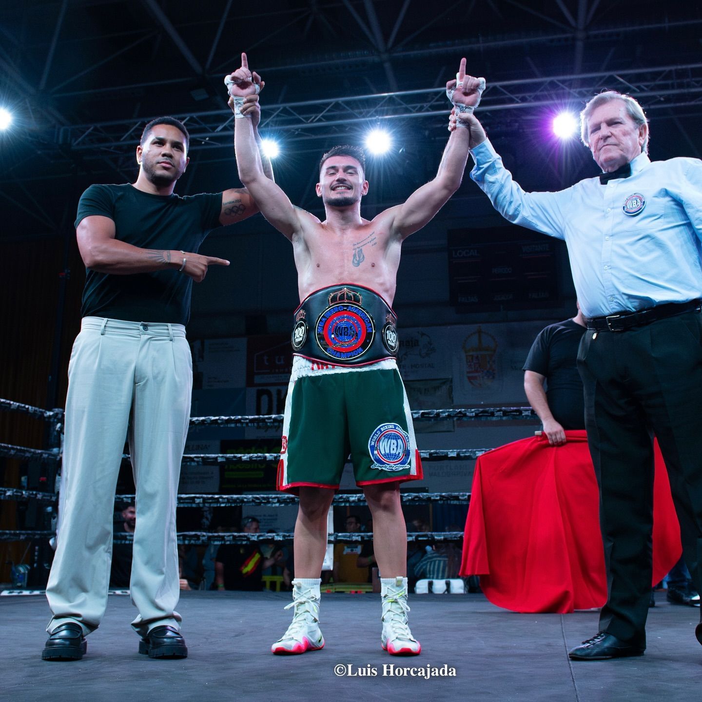 Boxeador en pantalones cortos verdes celebra la victoria, con los brazos en alto, cinturón de campeonato, árbitro y entrenador.