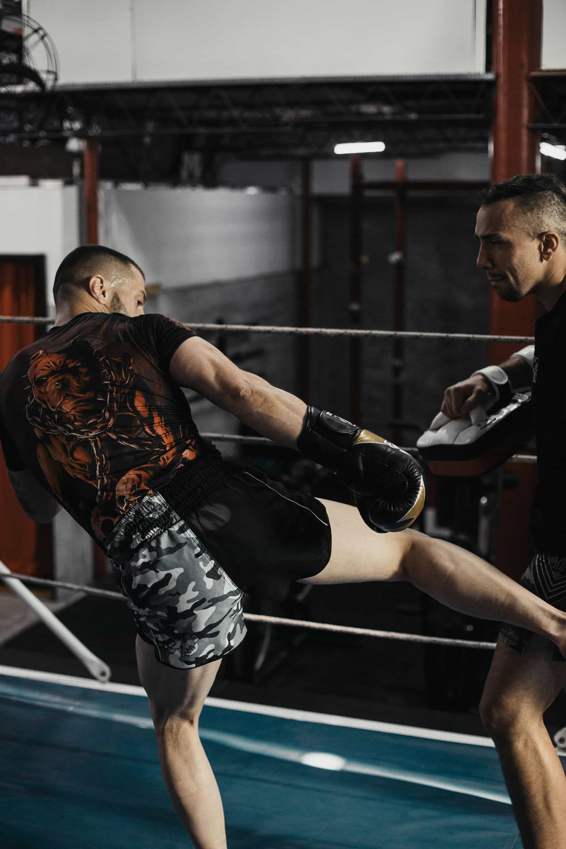 Un hombre patea a otro en un ring de boxeo. Ambos llevan ropa deportiva y guantes oscuros.